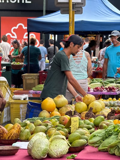 This image presents a bustling outdoor market scene in Papeete, France, under bright daylight. The foreground is dominated by extensive displays of fresh produce arranged on tables covered with red fabric. Prominently featured are large pale-yellow and light-green pomelos, mounds of green mangoes, several striped yellow and brown cacao pods, two heads of light green cabbage, green beans, and piles of dark, lumpy root vegetables. A small yellow sign on the table reads "Mangues 500F".

A young adult male stands centrally in the mid-ground, leaning forward over a produce stall. He has dark hair, a light mustache, and wears a dark green t-shirt and a red and black baseball cap reversed. A white bracelet is visible on his right wrist. Behind him, partially visible, is a female figure wearing a light blue lace-patterned tank top with a distinct criss-cross back design, and a tattoo is discernible on her right upper arm. To the right, another adult male wears a grey baseball cap and a light blue t-shirt; he is looking left and holding a small object in his right hand.

In the mid-ground, additional market tables display rows of green cucumbers, a pile of small red fruits, and lemons or limes. Blue market canopies extend across the upper portion of the frame, providing shade. Various plastic crates in yellow, blue, and red are stacked behind the produce stalls. Numerous other individuals, appearing to be both shoppers and vendors, are visible throughout the background and mid-ground. A yellow metal pole, likely part of a street fixture, stands centrally in the mid-ground. In the distant background on the left, a dark building features a large red section with a white graphic logo, partially readable as "PXP".
