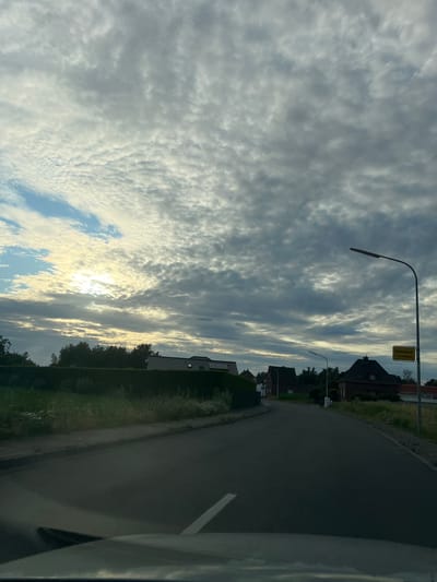 The image shows a road winding through a suburban area with houses and trees lining the sides. The sky is filled with dramatic clouds, with the sun peeking through, suggesting it is either sunrise or sunset. The lighting creates a somewhat muted and serene atmosphere. 

The image is oriented vertically, with the road and houses appearing on the right side of the frame and the sky dominating the left. A street light is visible near a road sign that reads "Unterbrech" followed by what appears to be a town name. The road itself is asphalt, with a white line marking its edge. The houses are of a traditional European architectural style, with pitched roofs. Greenery, including grass and trees, is abundant, indicating a rural or semi-rural setting. The overall impression is of a quiet, suburban landscape captured during a transitional time of day. There are no people visible in the image.