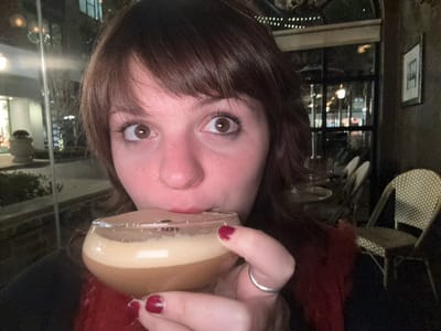 A woman with brown hair and eyes holds a glass of a light brown drink to her lips. She gazes forward with wide eyes. Her nails are painted a dark red. A silver ring adorns her finger. She wears a red, fuzzy garment. The background includes restaurant furniture, a framed picture on the wall, and a window showcasing outdoor lights. The entire image is oriented upside down.