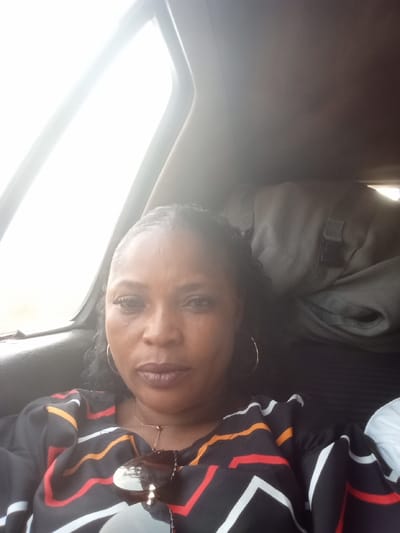 A selfie-style portrait depicts an adult female seated within the passenger compartment of a vehicle, reported to be in Itobe, Nigeria. The subject, with dark skin and dark braided hair, maintains a direct gaze towards the camera, exhibiting a neutral facial expression. She is adorned with large gold-colored hoop earrings and a thin gold-colored necklace. Her attire consists of a black garment featuring prominent red, orange, and white linear patterns. Dark sunglasses rest on her upper chest. To the subject's upper right and rear, a light-colored, possibly olive green, soft-sided bag with visible buckles is partially present. The vehicle's interior appears dark, contrasting with an overexposed window on the left side of the frame, indicating bright outdoor light.