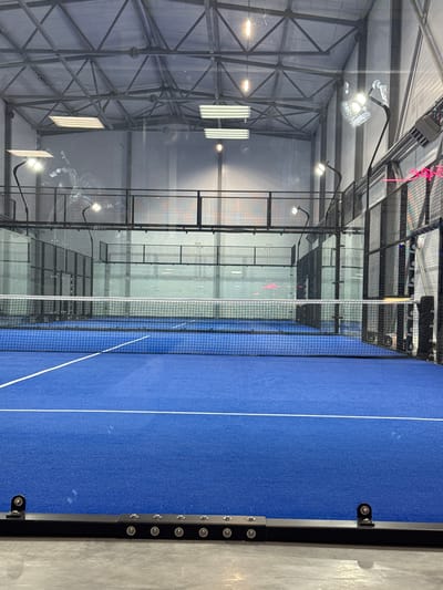 This media file shows an indoor Padel tennis court in Lubnya, Russia. The court features a vibrant blue surface with white lines marking the boundaries. A black net divides the court in half. The court is enclosed by glass walls and a metal mesh structure, allowing visibility into the surrounding facility. Several bright LED lights are mounted on the ceiling, illuminating the court, suggesting it is either daytime or the facility is lit for evening play. The overall scene depicts an empty sports venue, indicating no activity is currently taking place. The metallic structure of the building and the modern design of the court suggest a contemporary indoor sports complex. There are no people visible in the frame. The location cues are the name "Lubnya, Russia" provided as context. There is no discernible text in the image.