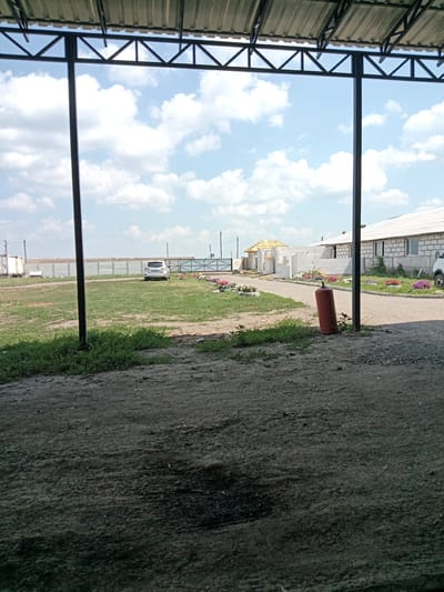 This media file shows a view from under a canopy or shelter, looking out onto an outdoor scene. The sky is bright blue with fluffy white clouds, suggesting it is daytime with fair weather.

In the middle ground, a white SUV is parked on a grassy area. Beyond the grass, there's a road and several buildings under construction, made of white brick and concrete blocks. Some areas are decorated with flowerbeds containing colorful flowers. A yellow-roofed structure, possibly a gazebo or small building, is also visible. A tall, orange gas cylinder stands upright near the foreground on the right side of the image. A fence runs across the background, separating the property from an open field or further industrial area.

The overall setting appears to be a rural or semi-rural location, possibly an industrial or construction site, with evidence of ongoing development. There is no visible text in the image. The scene is devoid of people, giving it a quiet and still atmosphere. The lighting suggests it is a sunny day, likely late morning or afternoon.