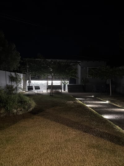This is a nighttime, exterior, low-angle shot of a modern building with a grassy area and outdoor seating. The scene is illuminated by artificial lighting, with spotlights highlighting a path alongside the grass and lights integrated into the building's facade.

The building appears to be made of concrete and glass, with several windows visible. A balcony or terrace area features outdoor furniture, including chairs and a table. A tree with dense foliage stands near the building, and smaller plants are present along the base of the structure and in the foreground. The grass area is sloped, with distinct rows of lighting embedded along its edge, creating a striking visual effect.

The time of day is clearly night due to the deep darkness of the sky and the reliance on artificial illumination. The weather appears to be clear, as there are no visible signs of rain or fog. The location in Nea Potidea, Greece, suggests a possibly Mediterranean setting, though no specific geographical landmarks are immediately apparent in the image. The overall mood is serene and contemporary, emphasizing architectural design and outdoor ambiance. No people or text are visible in the frame.