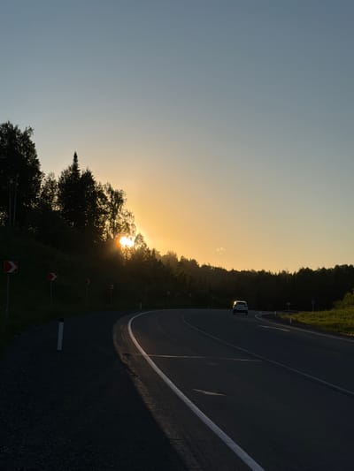 The image depicts a paved, two-lane road curving to the right, bordered by a grassy shoulder on the right and a tree-lined slope on the left. The sun is setting, casting a warm glow and silhouetting the trees. The sky transitions from a clear, light blue at the top to a soft orange and yellow near the horizon. The sun's rays pierce through the foliage, creating a bright focal point. A white car is visible driving on the road in the distance. Several red and white directional signs are placed along the left side of the road, indicating a curve. The scene is set in a natural environment, likely in or near the city of Kaz, Russia, given the visual context of a highway winding through a forested area during sunset.