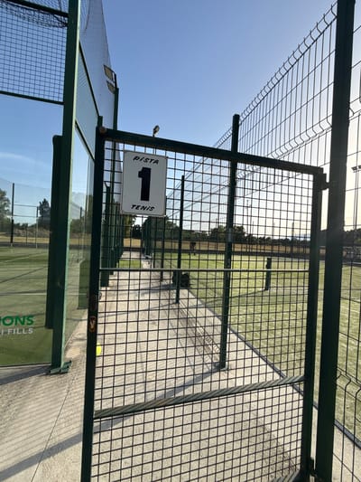 The image shows a fenced-off tennis court in Vidreres, Spain. A sign on the gate reads "PISTA 1 TENIS", indicating it is Court 1 for tennis. The court is made of green artificial turf, and a net is visible. The sky is clear and blue, with the sun setting in the background, casting long shadows. The scene appears to be a sports facility, possibly a public park or a private club, with other courts visible in the distance. There are no people in the immediate foreground of the image. The overall impression is one of a well-maintained outdoor sports venue on a sunny day.