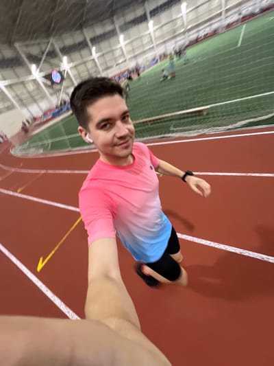 A selfie shows a man on a track. He wears a pink and light blue shirt, black shorts, and a smartwatch. He seems to be running. The track is red with white lines. In the background, there's a green field with a net and spectators. The venue appears to be an indoor sports facility.