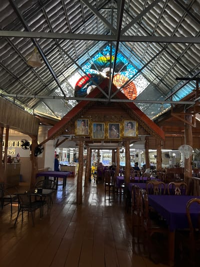 The image shows the interior of a restaurant in Tambon Ko Panyi, Thailand. The restaurant has a wooden floor and a high, sloped roof with exposed beams. The roof has a section of stained glass that allows natural light to filter in, with colorful fan-shaped designs visible. Four framed portraits of what appear to be royal figures hang on a wooden structure above a dining area. Several tables with purple tablecloths are set with chairs, and some tables are occupied by people. In the foreground, there are a few tables and chairs. Various ceiling fans and pendant lights are installed throughout the space. On the left side of the image, a sign is visible with English text that includes "UTE". The overall impression is of a well-lit, open-air dining establishment with traditional architectural elements and decorative features.