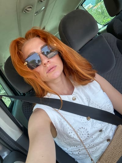 A woman with auburn hair and sunglasses is sitting in the passenger seat of a car, possibly in the city of Prijevorac, Montenegro. She is wearing a white eyelet top and a seatbelt. The interior of the car is visible, including the headrests and dashboard. Outside the car, green trees and houses can be seen. The overall lighting suggests it is daytime.