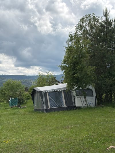 The image depicts an outdoor camping scene in Tsigov Chark, Bulgaria, under an overcast sky filled with dense, grey cumulus clouds that indicate diffused light. In the background, multiple layers of forested hills and mountains extend to the horizon, appearing hazy due to distance.
The midground features a white travel trailer or caravan, partially obscured by a slender deciduous tree, likely a birch, positioned in front of its rear section. Attached to the left side (viewer's perspective) of the caravan is a large, dark grey and black annex tent or awning, which has multiple transparent window panels and a lighter-colored roof section. To the right of the caravan and behind the birch tree, a dense cluster of tall, dark green coniferous trees stands.
Further to the left in the midground, a smaller, open-sided green canvas structure, possibly a changing tent or portable shelter, is erected on a visible wooden pallet base. Nearby on the ground, some dark, indistinct objects, possibly firewood or equipment, are present.
The foreground consists of an unmanicured grassy field, exhibiting various shades of green, interspersed with numerous small white clover flowers and occasional yellow wildflowers. No individuals or animals are visible in the scene.