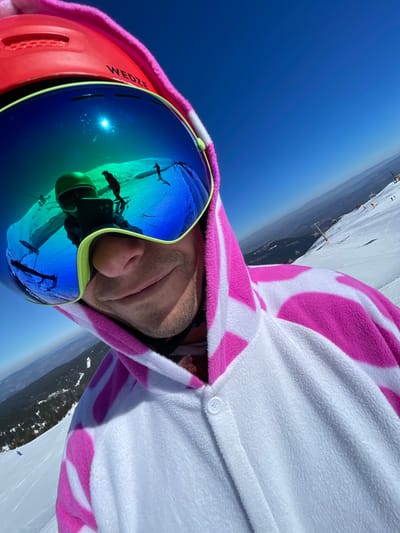 A close-up selfie captures an individual on a snow-covered ski slope in Jahorina, Bosnia and Herzegovina, under a clear blue sky. The subject wears a red ski helmet, branded "WEDZE," partially obscured by the hood of a white fleece one-piece ski suit featuring irregular pink markings. Large, reflective blue-purple mirrored ski goggles with a lime green frame cover the eyes. The goggles display reflections of a bright sun, a clear blue sky, a figure (likely the photographer) wearing a green helmet and holding a phone, and at least two additional figures skiing or snowboarding on a distant snow slope. Below the goggles, the individual's nose, mouth, and chin, showing light facial hair and a slight smile, are visible. The foreground reveals the white fleece material and white buttons of the ski suit. The background features expansive snow-covered mountain terrain, visible ski lift pylons on the right, and distant mountain ranges under a clear, bright sky. The image's perspective is tilted, consistent with a handheld selfie taken on a slope.