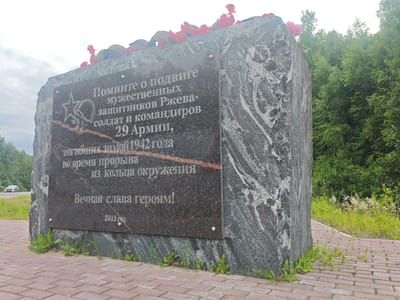 An outdoor, medium shot captures a granite monument with a plaque inscribed in Russian, situated on a brick-paved walkway in Esinka, Russia. The monument is topped with several Soviet-era military helmets and artificial red flowers. The plaque reads, "Remember the feat of the courageous defenders of Rzhev - soldiers and commanders of the 29th Army, who perished in the winter of 1942 during the breakthrough from the encirclement. Eternal glory to the heroes! 2013." The background features a dense green forest and a cloudy sky. A dark-colored car is visible in the distance on a road to the left of the monument.