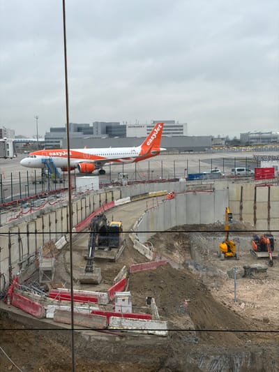 The image depicts an active construction site in the foreground with an airport apron and aircraft visible in the background, located at Charles de Gaulle Airport (CDG) in Tremblay-en-France, France, under an overcast sky.
In the mid-ground, a white and orange EasyJet Airbus A320 series aircraft is parked on an asphalt apron. A blue mobile air stair is connected to the forward left door of the aircraft. Two individuals, possibly ground crew, are discernible near the base of the air stair. In the background, airport terminal buildings, including one with "AIRFRANCE" visible, and other airport infrastructure are present.
The foreground is dominated by a large, multi-level construction excavation. A curved concrete retaining wall, topped with safety barriers, forms a pathway or access road descending into the pit. A large black and yellow excavator with a digging bucket is positioned on this curved pathway. Further into the excavation on the right, a yellow aerial work platform and a smaller orange excavator with a hydraulic breaker attachment are visible. Piles of excavated earth are scattered throughout the site. Concrete barriers, red and white construction barriers, and metal scaffolding are present around the excavation edges and within the pit. Three construction workers wearing high-visibility orange clothing are visible on the upper concrete retaining walls, two to the right and one closer to the center, appearing to be engaged in work or observation. A vertical metal pole in the center foreground obscures a small portion of the view.