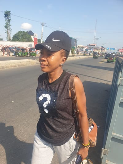 A woman wearing a black Nike baseball cap, a black tank top with white sequin accents, and light gray sweatpants stands on the side of a road in Aba, Abia, Nigeria. She has a brown handbag slung over her shoulder and is holding a blue mobile phone. The background includes a paved road, buildings, and several green tricycles ("keke") a common form of transportation in the city. Overhead power lines and a tall antenna tower are visible. The time of day appears to be sunny daylight.