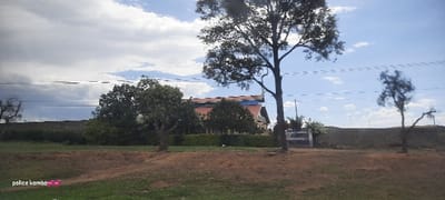A building with a red roof and gray walls is situated behind a green hedge and several trees. A gray stone wall extends to the sides, interrupted by a gate on the right. Power lines stretch horizontally across the sky, which is filled with clouds. A dirt bank with some green vegetation forms the foreground. Trees frame the view on the left and right.