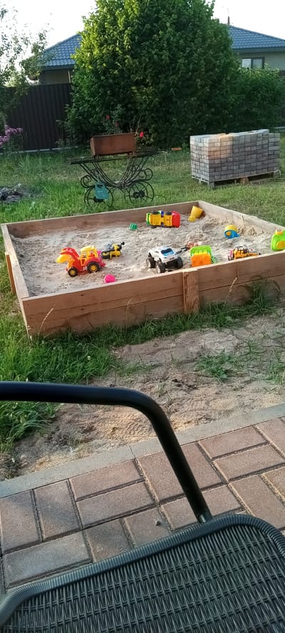 The image depicts a sunny outdoor scene in Hantsavichy, Belarus, likely during the late afternoon given the warm lighting and shadows. The primary focus is a wooden sandbox filled with sand and an assortment of colorful toy construction vehicles, including a red and yellow digger, a yellow motorcycle, and a white police truck with large wheels. A yellow toy dump truck lies overturned in the sand, alongside a yellow bucket and a pink ball. Scattered green and yellow plastic toys are also visible.
The sandbox is situated on a patch of green grass. In the background, a dark brown wooden fence runs along the property line, partially obscured by lush green trees and bushes. A house with a dark grey roof and some windows is visible behind the trees. To the right of the sandbox, a stack of grey and brown paving stones rests on a wooden pallet. A decorative, wrought-iron garden ornament with a rusty metal planter sits on the grass to the left of the sandbox, near some pink flowers.
In the foreground, the edge of a black metal chair arm and a portion of a dark wicker-patterned chair seat are visible, suggesting the photo was taken from a seated perspective, perhaps from a patio or seating area. The ground beneath the chair is paved with reddish-brown brick pavers. The overall impression is a relaxed, family-friendly backyard setting designed for outdoor play. No individuals are present in the frame.