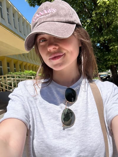 A young woman wearing a grey baseball cap with pink writing, a light grey t-shirt, and sunglasses is positioned in front of a multi-story yellow building with rows of windows. The building appears to be in Siófok, Hungary. A strap, possibly from a bag, is visible across her chest. Trees are visible in the background. She is sticking out her tongue slightly.