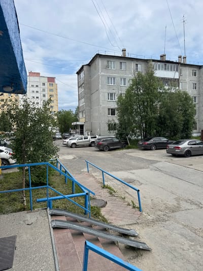 The image depicts a residential courtyard in Usinsk, Russia, on a cloudy day. In the foreground, a blue metal handrail and ramp system leads up to a building entrance. Several cars are parked in the courtyard, including a silver pickup truck, a dark sedan, and a silver sedan. Behind the parked cars, a multi-story apartment building, typical of Soviet-era architecture, dominates the scene. The building is constructed from concrete panels and features numerous windows. Trees and other vegetation are present, indicating a moderately green environment. The sky is overcast with scattered clouds.