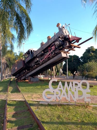 This image depicts a public monument featuring a black steam locomotive and its tender displayed on an elevated, angled metal structure, giving the impression of the train tipping upwards. The train, bearing the number "44" on its cab and a "NRB" logo on its tender, is positioned over a grassy area with actual railway tracks curving into the foreground on the left. In the bottom right foreground, large white letters spell out "CAMPO GRANDE," confirming the location as Campo Grande, Brazil. The scene is set under a clear blue sky with bright sunlight, casting shadows. To the left, a row of tall palm trees stands, and in the background to the right, various deciduous trees and a lamppost are visible. A single individual is discernible in the distant right background near the trees.