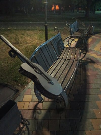 An acoustic guitar rests on a metal and wooden park bench in Biaroza, Belarus. The bench is positioned on a paved walkway with colorful interlocking bricks. To the left of the bench, a metal trash receptacle is visible. In the background, a lamppost stands beside a grassy area. The scene is dimly lit, suggesting it is nighttime or dusk, with shadows cast by the bench and guitar onto the walkway. The background is blurred, showing trees and foliage.