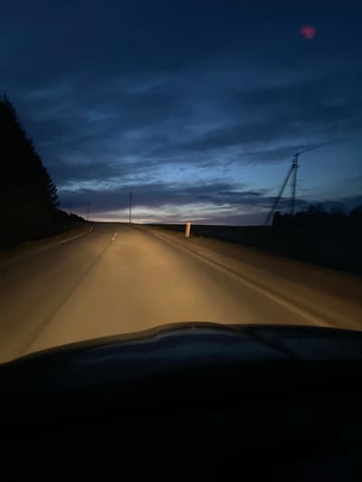 The image captures a twilight or nighttime scene from the interior of a vehicle, looking out onto a road in Kamennyi Kliuch, Russia. The sky, a deep indigo blue with soft, streaky cloud formations, dominates the upper-left and left side of the frame. A small, distinct red light, possibly a distant tower light or an external reflection, glows in the upper-left corner of the sky.

The road occupies the right side of the image, brightly illuminated by a warm, yellowish light, suggesting the vehicle's headlights. It appears to curve gently as it recedes into the distance, meeting a dark, indistinct horizon line formed by trees or low hills. Several slender utility poles or communication towers are silhouetted against the lighter part of the sky, with one pole featuring a prominent triangular support structure.

The extreme right side and lower portion of the image are very dark and out of focus, indicating the presence of the vehicle's interior, such as a dashboard or door frame. No people are visible, nor is any discernible text. The overall scene evokes a sense of tranquil travel through a rural or semi-rural landscape during the early morning or late evening hours.