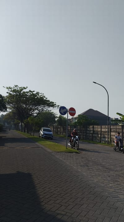 The image depicts a street scene in Sidodadi, Indonesia, under a clear, bright sky. The foreground is dominated by a paved road, likely made of interlocking bricks, with a distinct shadow cast across it from the left. A grassy median runs along the right side of the road.
On the right side of the paved area, two individuals are riding motorcycles. The first motorcycle, closer to the viewer, carries two people; the rider is wearing a dark-colored shirt and a head covering, and the passenger is less clearly visible. The second motorcycle, further down the road, has a single rider wearing a light-colored shirt.
A tall, curved streetlamp stands on the right side of the road, extending into the sky. Two traffic signs are mounted on a single pole. The upper sign is a blue circle with a white arrow pointing left, indicating a mandatory direction. The lower sign is a red circle with a white horizontal bar, signifying a "no entry" or similar prohibition.
To the left of the motorcycles and signs, a silver car is parked on the side of the road, facing away from the viewer. Beyond the car, a row of trees with dense foliage lines the left side of the road, extending into the distance. A concrete wall, likely a boundary for properties, runs along the right side of the road, separating it from buildings that are partially visible behind it. The architectural style of these buildings suggests a residential area. The overall impression is a quiet, possibly suburban street in the morning or late afternoon, given the quality of light and the presence of shadows.
