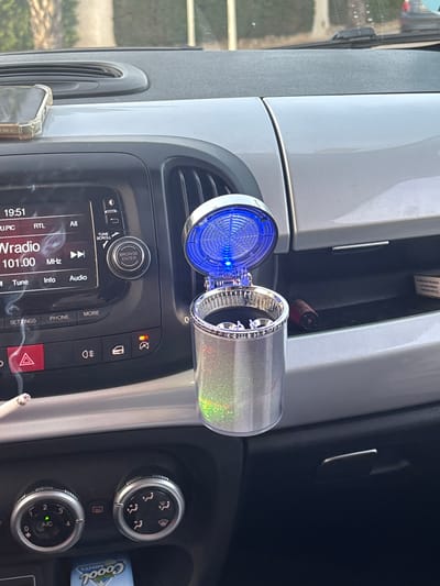 In Urbanización Cabo Roig, Spain, a silver car ashtray with a blue LED light and glittery exterior is placed in the center console of a car. The car's radio display shows "19:51" and "Wradio 101.00 MHz". A lit cigarette is visible in the foreground, next to the dashboard. Various car controls, including hazard lights and air conditioning settings, are also present. In the background, a pack of cigarettes and a red cylindrical object, possibly a lighter or vape, are visible in a storage compartment. The dashboard and surrounding interior are light gray and black.