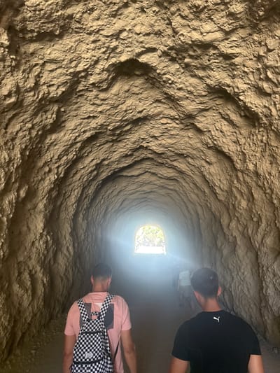 The image shows the interior of a rough-hewn tunnel, possibly a cave or an ancient man-made passage. The walls are textured with natural, earthy tones, creating a sense of depth and mystery. Sunlight streams in from an opening at the far end of the tunnel, illuminating the space and creating a bright contrast with the darker interior.

Two individuals are visible in the tunnel, walking away from the camera towards the light. The person in the foreground is wearing a pink t-shirt and a black and white checkered backpack. The person behind them is wearing a black t-shirt with a white logo. Their presence suggests exploration or a visit to this unique location.

The scene appears to be set during the daytime, indicated by the bright sunlight. The overall mood is one of adventure and discovery, with the natural rock formations and the distant light inviting further exploration. There is no visible text in the image. Given the location context of Ardales, Spain, this could be an archaeological site or a natural geological formation.