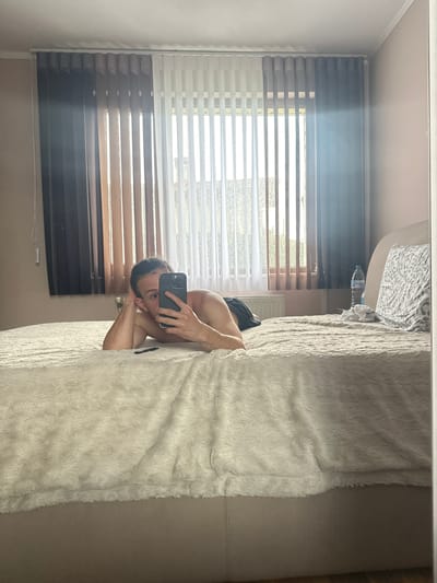 The image depicts an indoor scene in a bedroom located in the city of Saraia, Bulgaria. The photograph is a selfie taken in a mirror by a person, appearing male, lying prone on a large bed covered with a light-colored, textured faux fur or fleece blanket. The individual's torso is bare, and dark shorts are visible. Their left arm supports their head, while their right hand holds a dark-colored smartphone, obscuring most of their face except for their left eye, nose, and forehead. A black pen-like object lies on the bed in front of them.
Behind the bed, a large window spans the wall, fitted with vertical blinds or curtains. These coverings transition in color from dark brown/black on the left, to medium brown/tan in the center, and white/translucent on the right. Bright sunlight streams through the partially open blinds, creating strong highlights and a subtle lens flare in the upper right quadrant of the image.
To the right of the person, a light beige headboard is visible, against which a pillow with a grey and white patterned case rests. A clear plastic water bottle with a blue cap and a crumpled pile of white fabric are positioned on the bed near the headboard. A white radiator is visible below the window on the far right. The walls are a light, possibly beige or pinkish-beige, and the ceiling is white with molding.