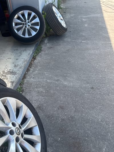 The image shows four car wheels and tires placed on a concrete surface in Schkopau, Germany. The wheels appear to be alloy, with a multi-spoke design. Three of the wheels are visible in their entirety, while a fourth is partially cropped at the bottom of the frame. The tires are black and appear to be in good condition. The concrete surface is gray and shows signs of wear and tear, with some patches of dirt and debris. The background is out of focus, but it appears to be an outdoor setting, possibly a driveway or a garage area. The lighting suggests it is daytime, with shadows cast by an unseen object on the right side of the frame.