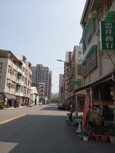 The image shows a street scene in Kaohsiung, Taiwan, with a focus on the quiet, sunny day. The street is lined with a mix of low-rise and high-rise buildings, including apartments and commercial establishments. On the right, a building displays a green sign with Chinese characters, and a street sign reads "Gueisuei St." A teal scooter is parked along the sidewalk amidst potted plants and a vendor's sign. A red car is parked further down the street. In the distance, traffic signals and vehicles can be seen. The asphalt road has double yellow lines. The sky is clear and pale blue.