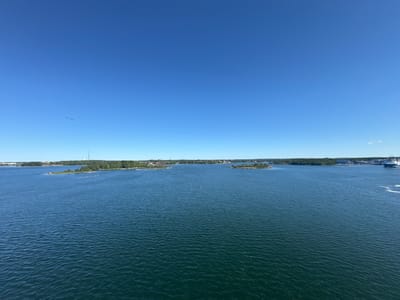 The image captures a wide view of a body of water with several islands dotting the horizon, set against a clear blue sky. The water is calm with gentle ripples, reflecting the bright sunlight. Lush green trees cover the islands, and some small structures, possibly houses or buildings, are visible on the mainland in the distance. A tall tower with an antenna stands prominently on one of the islands. In the far right of the image, a large blue and white ship is partially visible, moving away from the viewer. A single bird is seen flying in the upper left quadrant of the sky. The scene suggests a tranquil day in a coastal or archipelago setting. This view is consistent with the archipelago surrounding Karlskrona, Sweden, a city known for its naval history and its location within a scenic coastal landscape.