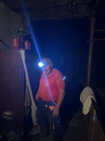 The image depicts a man wearing a red t-shirt and dark pants, with a headlamp illuminating his face, standing in what appears to be an outdoor or semi-outdoor area at night. The headlamp beam is bright white and casts a blueish hue on his face and the surrounding area. He has a smiling expression and is looking slightly to his left.
To his left, there are two potted plants on a shelf, one with lush green foliage in a reddish-brown pot, and another smaller plant behind it in a similar pot. To the far left, a dark, textured surface, possibly a wooden or corrugated metal wall, is visible. A white sheet or fabric hangs down from the ceiling area on the left side of the frame.
In the background, the scene is dark, with hints of foliage or vegetation visible. A vertical support structure, possibly made of metal, is present on the right side of the image. A folded, striped cloth or blanket is draped over a wooden surface on the far right. The ground appears to be uneven, possibly made of stone or dirt. The overall impression is that of a functional, perhaps slightly rustic, environment illuminated by the headlamp. The provided location of Tandzia, Georgia, suggests a rural or semi-rural setting within that region.