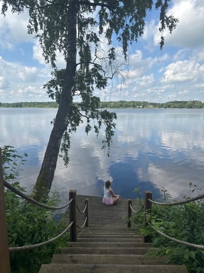The image depicts a serene waterside scene with a young person sitting on a wooden dock. The person is wearing a white tank top and pink shorts, with their legs crossed in a meditative pose, facing the expansive lake. The dock, constructed from weathered wooden planks, descends in a series of steps towards the water. Rope railings with wooden posts guide the way down the steps. Lush green vegetation, including ferns and other foliage, borders the dock, with a large, leaning tree with dappled leaves providing a natural canopy. The lake itself is calm and reflects the cloudy sky, creating a mirror-like surface. In the distance, a treeline marks the opposite shore, with hints of structures visible amongst the trees, suggesting the presence of a settlement or buildings near Priedesmuiža, Latvia. The sky is a mix of blue with fluffy white clouds, contributing to the tranquil atmosphere.