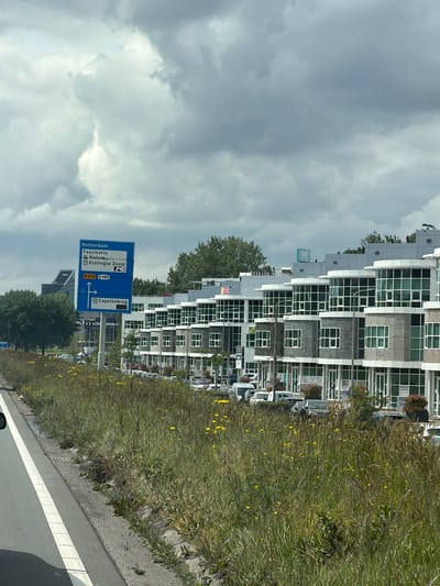 The image is taken from a roadway in Capelle aan den IJssel, Netherlands. The view is of a roadside with green vegetation and a series of modern, multi-story buildings with geometric facades. A blue highway sign indicates directions to Rotterdam, Fascinatio, Rivium, and Kralingse Zoom, as well as Capelsebrug via routes N210 and S107. The sky is overcast with clouds. The image appears to have been rotated counterclockwise for presentation.