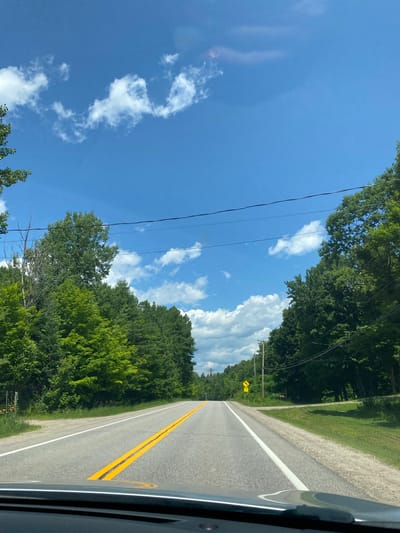 The image depicts a view from inside a vehicle driving on a paved, two-lane road bordered by dense green trees on both sides. The sky is bright blue with scattered white clouds. In the distance, a yellow diamond-shaped sign with the number "75" is visible on the right side of the road, indicating a speed limit. The road features a double yellow line in the center, separating the lanes. The foliage suggests a rural or semi-rural setting, likely during the daytime. The specific location in the city of Lac-Simon, Canada is not ascertainable from the visual information provided.