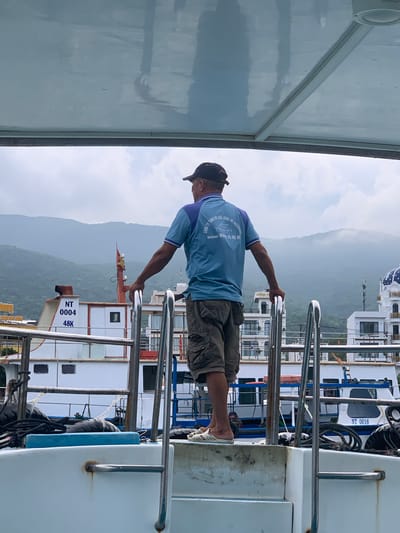 An adult male wearing a blue and purple polo shirt, khaki cargo shorts, a black baseball cap, and white slip-on shoes stands on the deck of a white boat, facing away from the camera. His hands are resting on the railing of the boat. In the background, there are other boats, buildings with white facades, and lush green mountains under a cloudy sky. The text on the man's shirt reads "CÔNG TY TNHH DU LỊCH VÀ LỮ HÀNH HY DISCOVERY" and "VINH HY", along with a phone number. The boat the man is standing on has "NT 0004 48K" written on its side. Another boat in the background has "NT 0016" visible. The overall scene suggests a harbor or coastal area in Vinh Hai, Vietnam.