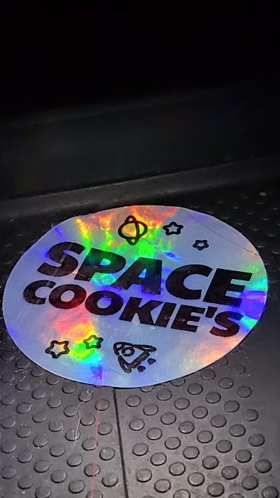 A circular holographic sticker is affixed to a dark, studded, rubberized floor surface in Magaluf, Spain. The sticker displays the black uppercase text "SPACE COOKIE'S," complemented by outline graphics including an atom-like symbol, multiple stars, and a rocket ship with exhaust trails. The holographic material reflects a full spectrum of rainbow colors. Minor creases and surface wear are visible on the sticker. The underlying floor is dark grey or black, characterized by a uniform pattern of raised circular studs, with a faint linear seam present to the right of the sticker. The upper portion of the image is indistinct and dark, indicating a low-light environment.