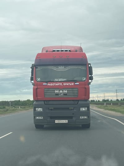 The image shows a red MAN truck driving on a paved road. The truck has a Russian flag and text that reads "РАБОТАЙТЕ, БРАТЬЯ!" (Work, brothers!). There are also two smaller stickers on the windshield. The truck is driving in the direction of the camera. The environment is a rural setting with a cloudy sky and vegetation on the sides of the road. The license plate of the truck is visible and reads "P 003 EE 64". The image was taken in the city of Zorinskii, Russia.
