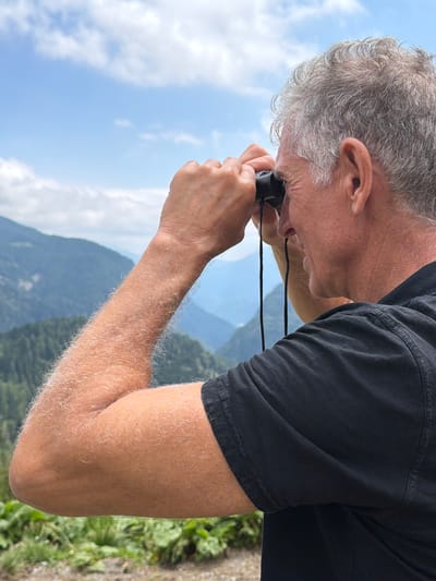 A middle-aged man with graying hair looks through black binoculars at a mountainous landscape. He is wearing a black t-shirt and his arms are tanned and covered in light hair. The background shows layers of green, forested mountains under a blue sky with scattered clouds. The scene is likely outdoors, in a natural setting like the Alps, possibly in or near Enemonzo, Italy, given the mountainous terrain.