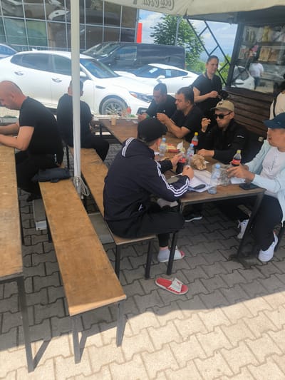 The image shows a group of men seated at a wooden picnic table outside a building, possibly a restaurant or store. The men are dressed in casual clothing, including black shirts, jackets, and baseball caps. Some are eating or drinking from bottles and paper wrappings. A woman in a black shirt stands behind them, possibly serving or supervising. The setting appears to be an outdoor patio area covered by a large white umbrella, casting a shadow on the ground. In the background, there are several parked white cars and a building with reflective windows. The ground is paved with patterned blocks. The scene is possibly located in Rustavi, Georgia, based on additional context.