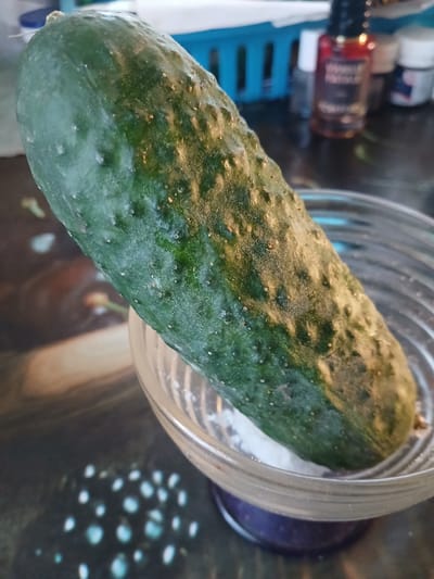 A close-up, eye-level shot focuses on a bumpy green cucumber resting diagonally across a clear glass bowl. The bowl itself appears to contain a white powdery substance, likely salt, and is positioned on a dark, reflective surface that displays abstract patterns of light. In the background, out of focus, are several bottles, including one with a red liquid, and a blue plastic basket. The lighting is dim, creating a moody atmosphere. The image does not provide any information about a location in Hlushky, Ukraine.