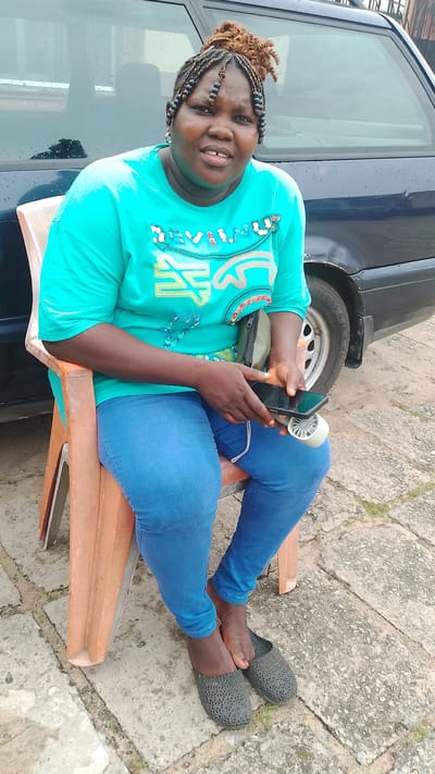 A middle-aged Black woman sits on an orange plastic chair on a concrete surface comprised of square tiles. She is positioned outdoors, next to a navy blue vehicle. She wears a turquoise T-shirt, blue jeans, and gray slip-on shoes. Her hair is styled in braids, with some sections pulled into a bun on top of her head. She holds a black smartphone and a small white portable fan. The location is Agbarho, Nigeria.