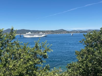 A wide-angle, eye-level shot captures a picturesque view of the Bay of Pampelonne, near Saint-Tropez, France, on a clear, sunny day. The foreground is dominated by lush green foliage, framing the scene from the bottom and right edges. In the middle ground, the turquoise waters of the Mediterranean Sea stretch out, dotted with several sailboats, including one large vessel with dark sails. Further in the distance, a large white cruise ship sails across the bay. Behind the sea, a hilly coastline is visible, covered with a dense network of buildings and vegetation, suggesting a coastal town. The sky is a vibrant, cloudless blue, with a faint, thin contrail visible in the upper portion of the frame. The overall impression is one of serene coastal beauty and leisure activity.