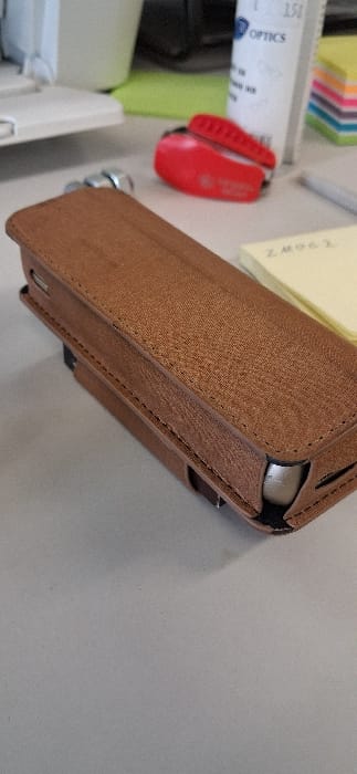 A close-up photograph captured in the city of Drugan, Bulgaria, depicts an organized desktop surface. In the foreground, a brown, textured folio-style case, likely designed for a mobile electronic device, rests horizontally on a light grey surface. The case exhibits visible perimeter stitching and incorporates metallic silver elements at its ends. In the mid-ground, positioned to the right of the case, a red object, consistent with a small folding utility knife, is present. Directly behind the red object, a white cylindrical container, partially labeled "OPTICS" with the numbers "150" visible, stands upright. Further to the right, a stack of multi-colored square sticky notes (pink, purple, blue, green) is visible, alongside a yellow sticky note bearing the alphanumeric text "ZMOG2". A grey writing instrument is positioned near the sticky notes. On the far left of the frame, a portion of a white electronic device, possibly an office machine, and a bright green object are discernible in the background. No individuals are present in the scene, and no overt actions or interactions are depicted.