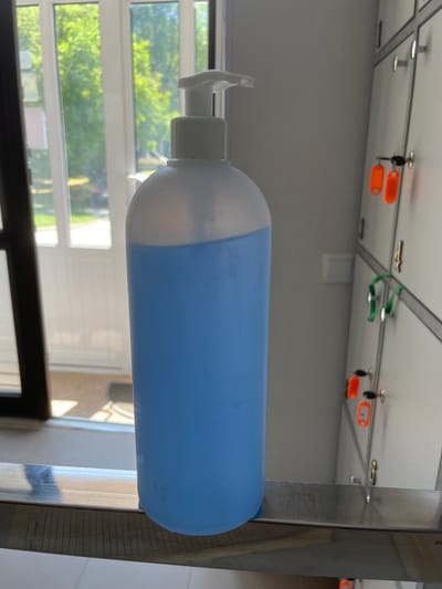 A tall, translucent white pump bottle filled with blue liquid stands on a metal shelf. The bottle is positioned in the foreground and is mostly in focus. In the background, a row of grey lockers is visible, some with orange key tags and green loops. Sunlight streams through a window to the left, illuminating trees and a doorway. The scene appears to be an indoor locker room or similar facility, possibly in Nisporeni, Moldova, given the contextual information provided. No people are present in the image.