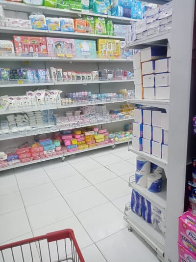 The image depicts an interior aisle of a retail store, situated in Nganjuk, Indonesia. The floor is composed of white square tiles with dark grey grout. A long, multi-tiered white metal shelving unit occupies the left side of the aisle, displaying various products. The top shelves stock packaged diapers and sanitary products, including brands like 'Lively' and 'Mamypoko'. Below these, baby care items such as 'Johnson's' gift sets, lotions, powders, and feeding bottles are arranged. The lower shelves feature numerous small, colored plastic containers in pink, yellow, blue, and green, along with other packaged goods. On the right, a narrower white metal shelving unit holds stacks of white tissue boxes on its upper shelves, and rolls of toilet paper, predominantly white with blue branding, on its lower shelves. Pink packaged items are visible at the bottom right of this unit. The red handle and upper wire basket of a shopping cart are visible in the lower-left foreground. No individuals are present in the frame, and no actions or interactions are depicted.