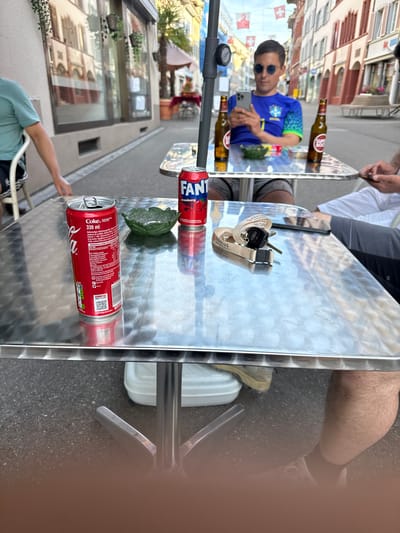 The image presents an outdoor café scene in Liestal, Switzerland, viewed from a high angle looking down at a highly reflective metallic table. The bright reflections suggest a sunny day.
On the table, various beverages are arranged: two brown glass bottles, one clearly labeled "SUPER BOCK," and three aluminum cans. One can is identified as "Fanta Strawberry," while two others are "Coke" (Coca-Cola), each 330 ml, with one open and the other closed. The "Coke" cans show text like "DOWNLOAD" next to a QR code and "100%". A dark green, leaf-shaped item, possibly a small dish, is also on the table. A set of car keys hangs from a lanyard bearing the word "ABACUS."
The table's reflective surface prominently mirrors a young man wearing sunglasses and a blue and green sports jersey, resembling a Brazil national team kit. He is holding a smartphone, possibly an iPhone. Partially visible are other individuals: an arm and leg of a person seated to the right of the table, and the arm and torso of another person in a light blue t-shirt, reaching towards the table from the lower left.
In the background, both directly and as reflections, are charming building facades decorated with multiple Swiss flags, confirming the given location. The ground is asphalt, and a white plastic container and parts of chair legs are visible beneath the table. The scene depicts a casual gathering at an outdoor establishment.