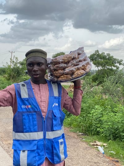 A young African man wearing a dark green cap and a pink traditional outfit stands on a dirt road in Rimin Ziem, Nigeria. He wears a bright blue vest with multiple pockets and reflective silver stripes. The vest has the word "Coupage" printed in white on the left chest. He carries a large, clear plastic bag filled with brown, oval-shaped items on his right shoulder. The background consists of lush green vegetation, trees, and a cloudy sky. A white plastic bottle is visible on the right side of the road.