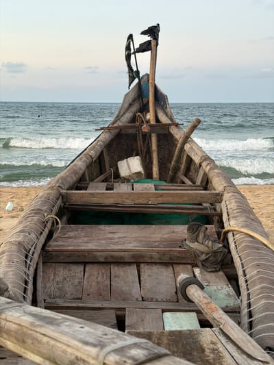 This is an outdoor shot of a rustic, wooden boat on the sandy shore of Thuận An Ward, Vietnam, facing the ocean. The frame is oriented vertically, showing the bow of the boat pointed towards the horizon. The boat is weathered, constructed from roughly hewn wooden planks and beams. It is fitted with various ropes, netting, and other implements, indicating it is a fishing vessel. The ocean's water is churning with small waves. The sky above the horizon is a pale, overcast gray.