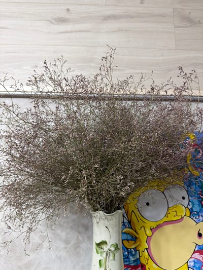 A bouquet of dried lavender is displayed in a decorative white ceramic vase. The vase features a hand-painted green clover motif. The arrangement is positioned in front of a colorful mural that depicts a cartoon character resembling Homer Simpson, with exaggerated features and dripping yellow frosting. The background consists of a light-colored wooden plank wall and a marble-patterned surface. The scene appears to be an indoor setting in Zarechny, Russia.